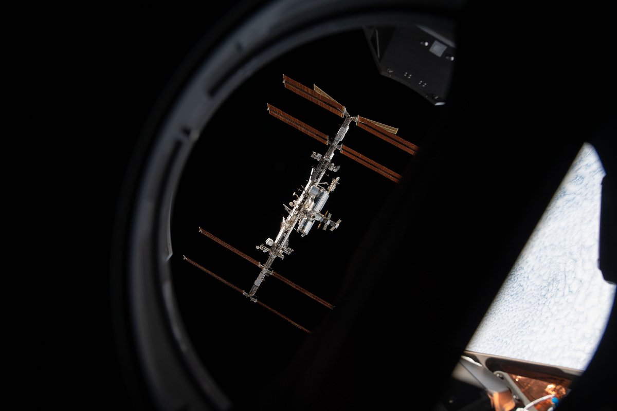 A photo of the International Space Station, seen in full against the black background of space, from inside the Crew Dragon capsule. The ring of the capsule's window is visible near the edges of the photo.