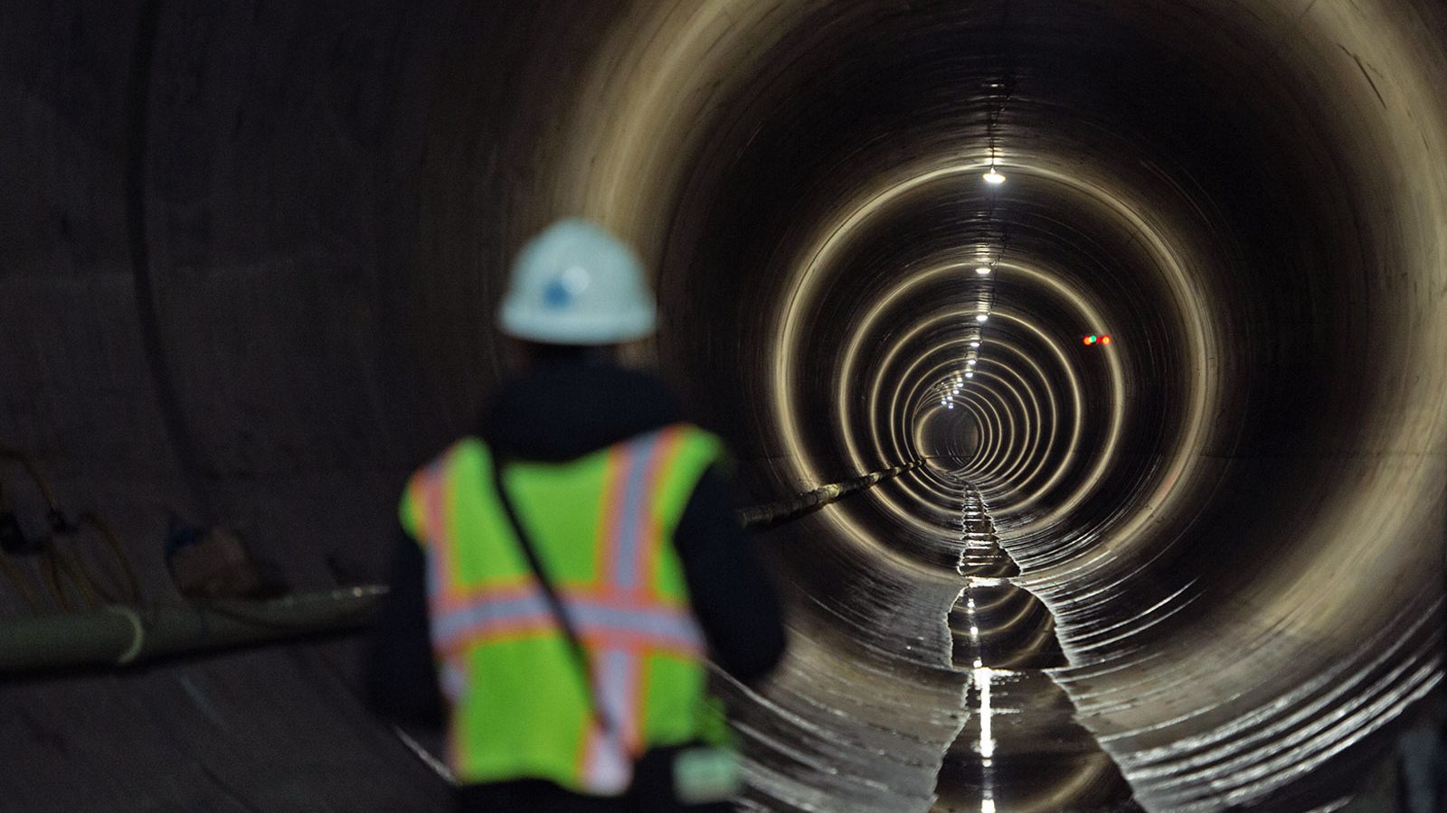 Rear view of person in high viz safety vest and white hard hat standing at entrance to large sewage tunnel