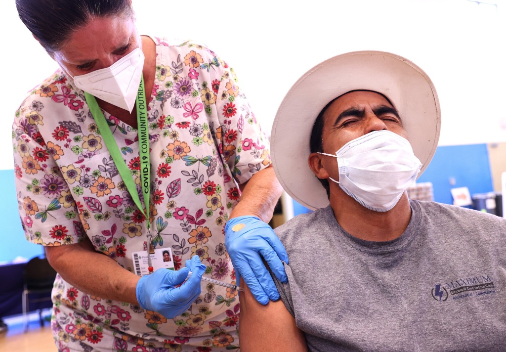 a person in a hat winces as a woman in blue gloves gieves him a shot