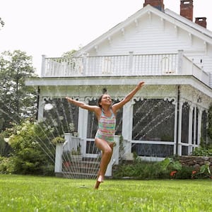 A girl running in the front yard while getting sprayed by sprinkler water