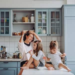 Laughing mom and two young daughters sitting on a kitchen island, eating a snack