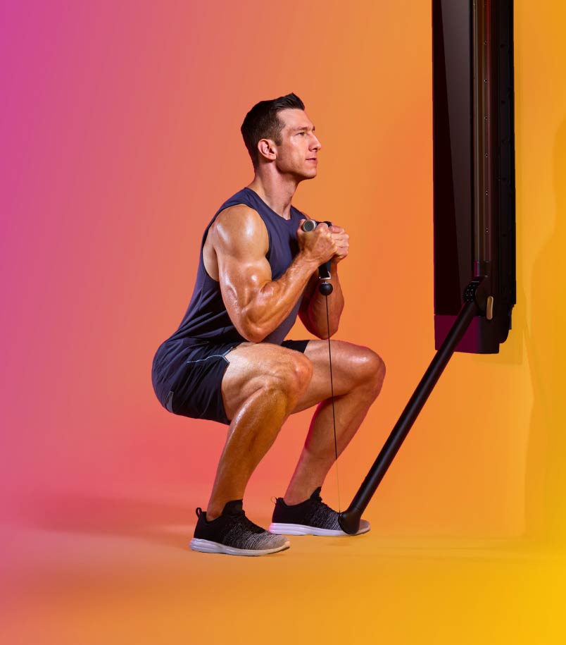 A man in workout clothes performs a lower body workout using a Tonal against a multi-colored backdrop.