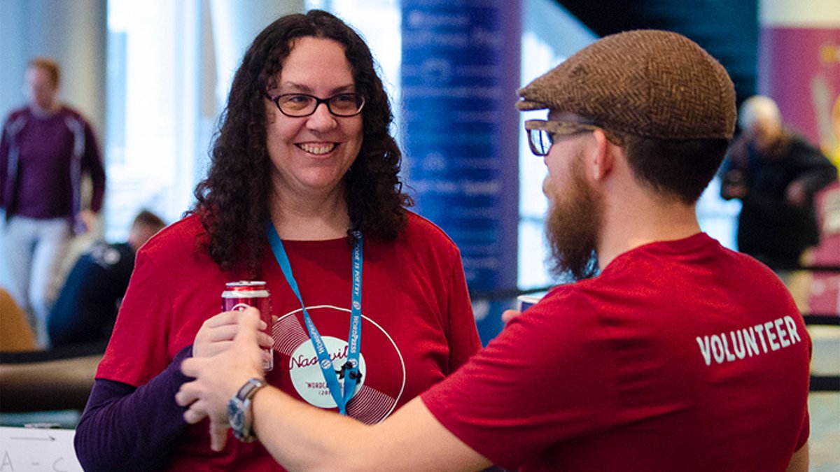 Two WordCamp US Volunteers talking. One is facing and smiling with the other holding his arms forward.