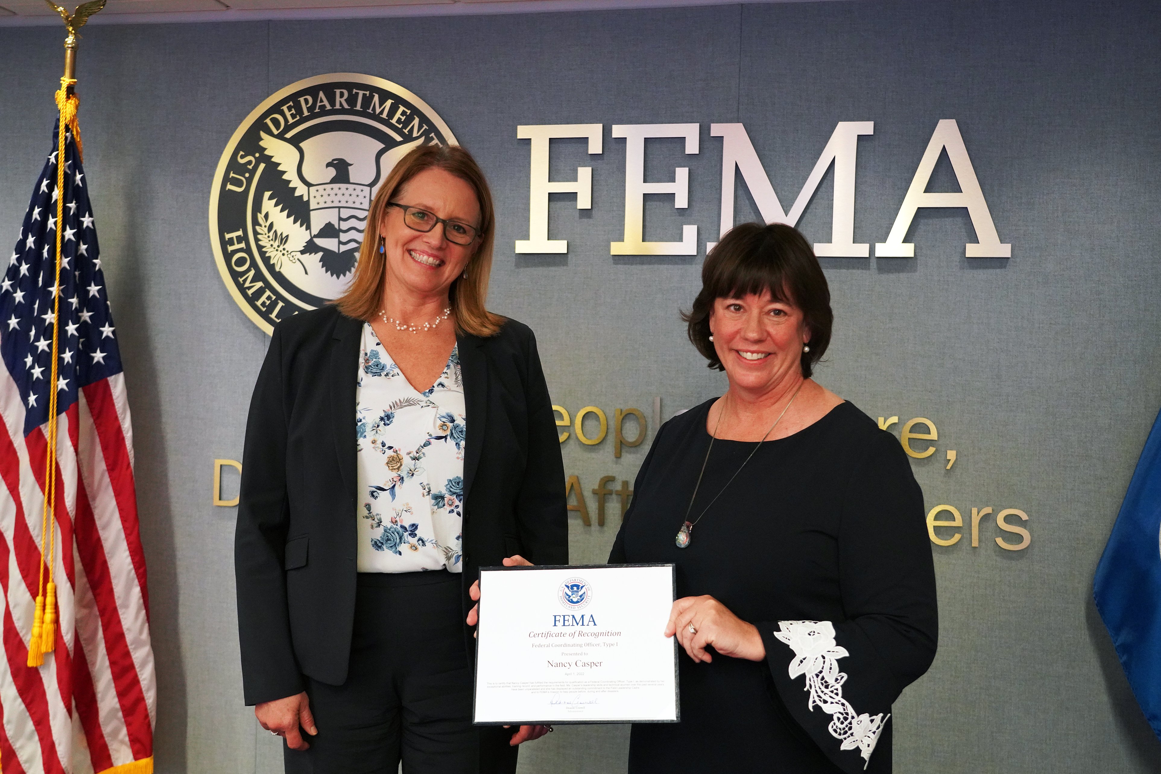 a photo of FEMA's Administrator Deanne Criswell standing by Federal Coordination Officer Nancy Casper who is holding a certificate in front of a wall with "FEMA" on the wall and an American flag to the left in the photo.