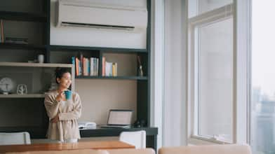 woman with cup of coffee near large windows and underneath air conditioning vent