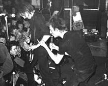 Two members of the rock band Crass are shown at a performance. From left to right are an electric guitarist and a singer. Both are dressed in all black clothing. The singer is making a hand gesture.