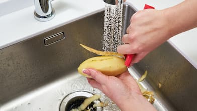 Hands peeling a potato over a garbage disposal while water runs from the faucet