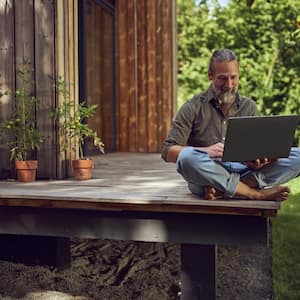 A mature man using a laptop while sitting outside a wooden house