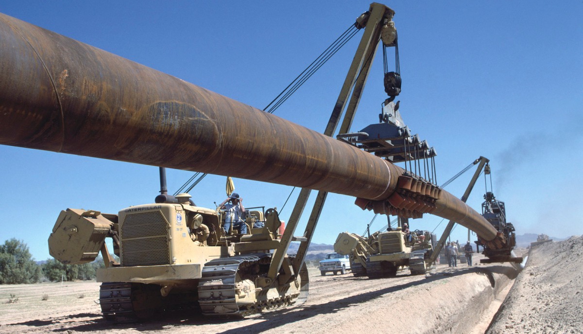 A crane lifts a section of pipeline into a trench