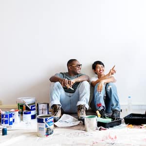 Couple sitting on floor surrounded by paint drop cloths, paint buckets, and a ladder