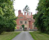 The side of a red-brick house with stone dressings seen along a drive. On each side of the drive is a lawn and the whole picture is framed by trees.