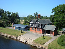 A small, two-storey, two-bay house seen from an elevated angle; the lower storey is in brick and the upper storey is timber-framed. To the right of the house is a low extension and to the left is a barn with a curved roof. In front of the house is a lawn and a slipway leading down to the river.