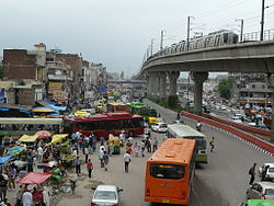 Red and orange coloured Delhi Transport Corporation Compressed Natural Gas buses amid pedestrian traffic below the elevated Delhi Metro train