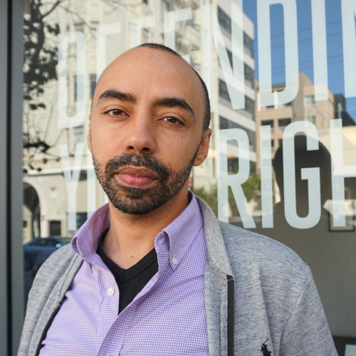 a photo of nash from mid-breast up, standing in front of a window with white laminate text spelling out "Defending Your Rights." 