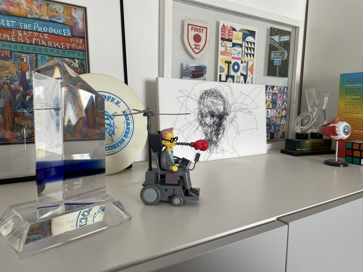 My 5  year Crystal alongside a simpsons Steven hawking character, an eyegaze painting of a somber face, a frisbee golf frisbee, a model of an Eyeball and Brad smiths tools and weapons book