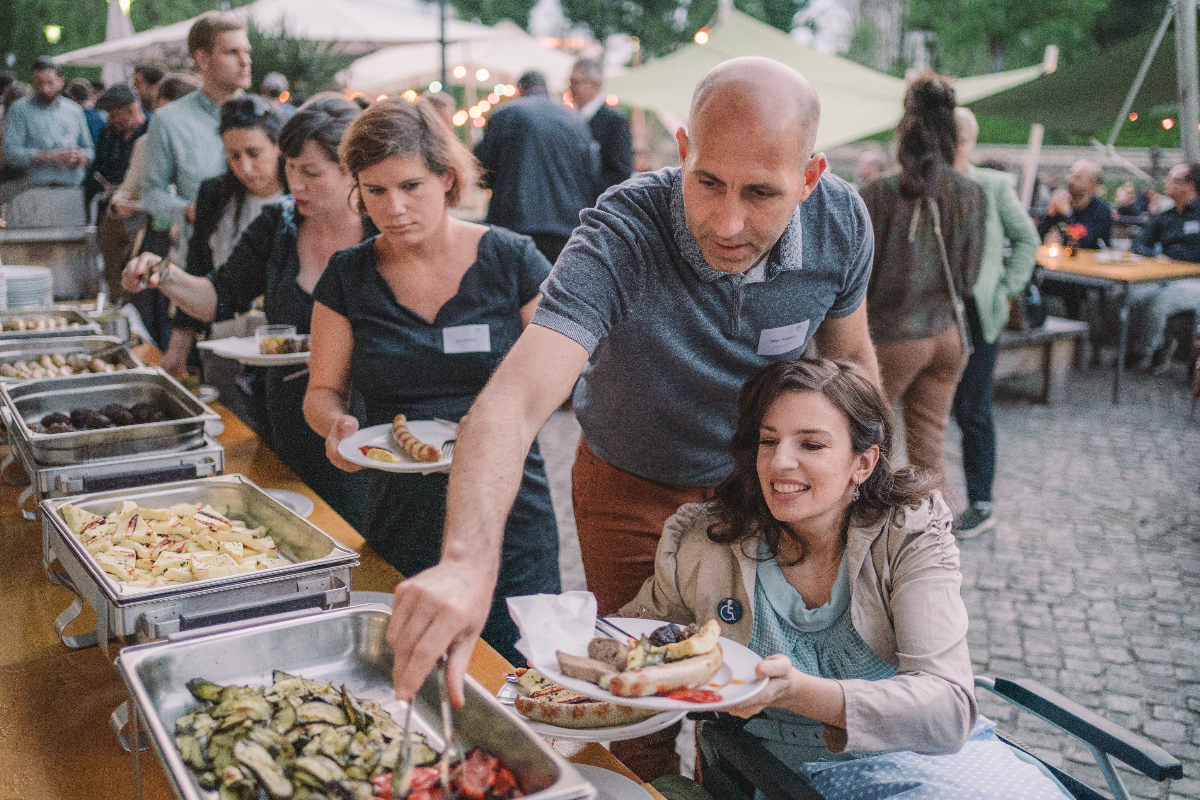 Dejan Mihajlovic gibt der sitzenden Maria Weisband Essen vom Buffet. Er steht dabei hinter ihr. Im Hintergrund ist eine Schlange am Buffet und eine  Ansammlung Menschen vor sommerlicher Terrasse erkennbar.