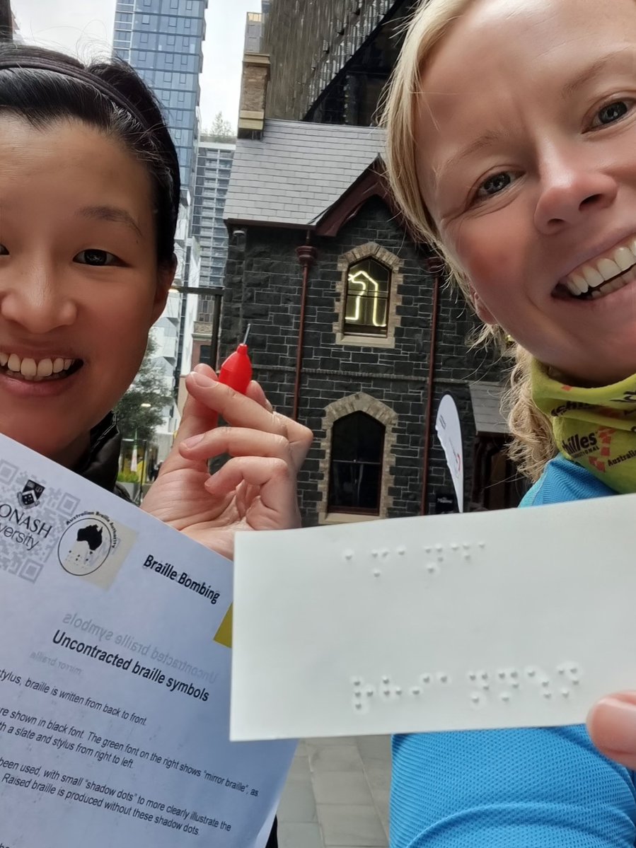 Selfie of two people (Rhi and me!) I am on the left holding my Braille reference sheet and Braille stylus. Rhi holds piece of sticky paper with our names printed in Braille. Dark bricked house-like building in the background.