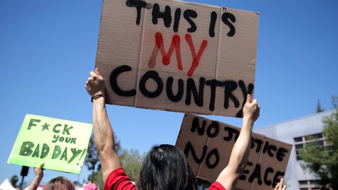 A person holds up a sign that reads "This is my country." Another person holds up a sign that says "Fuck your bad day."