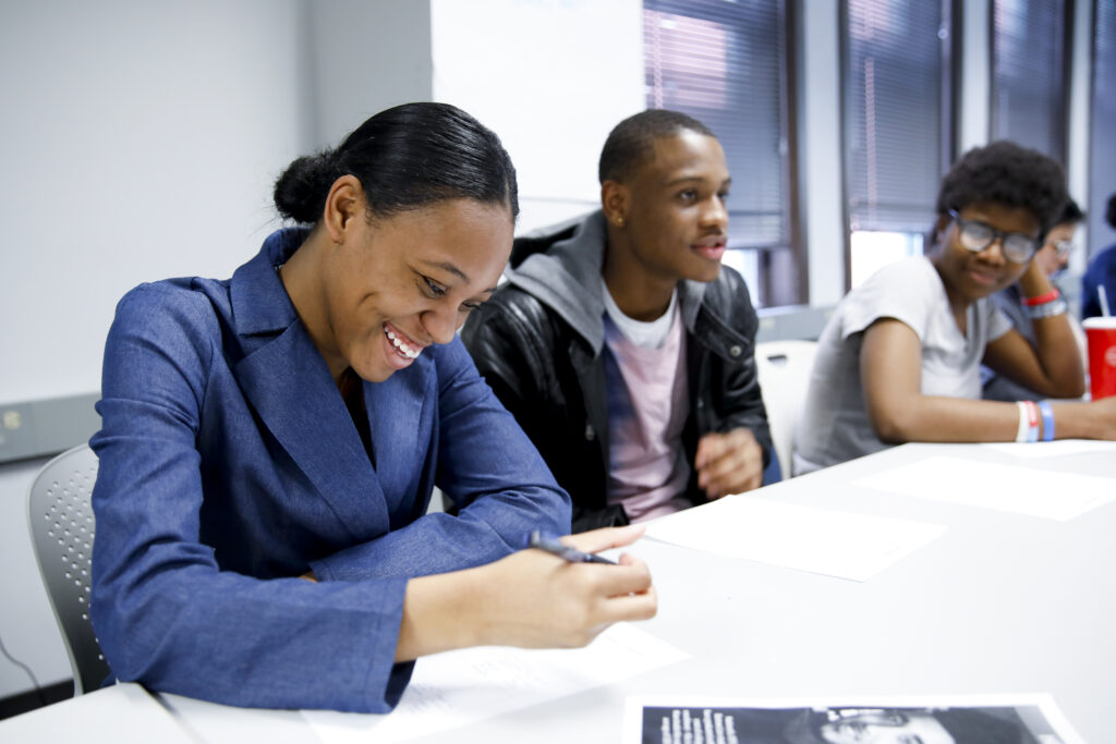 A young woman in a blue blazer smiles while writing on a piece of paper.