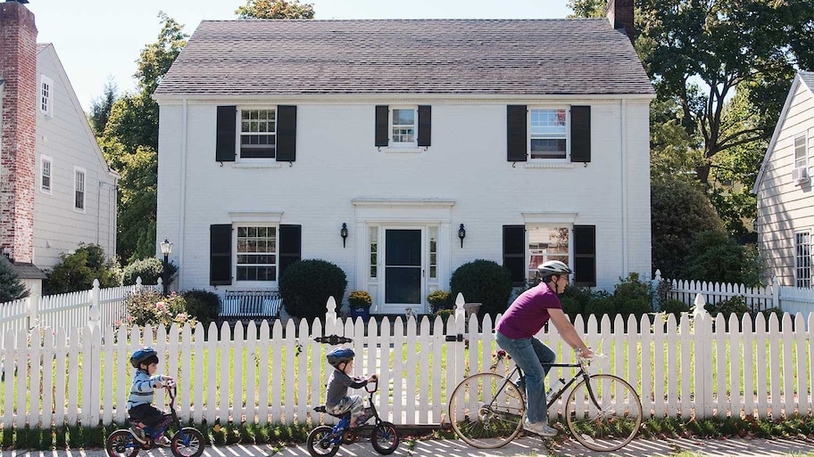 Father and kids riding bikes in front of fenced house