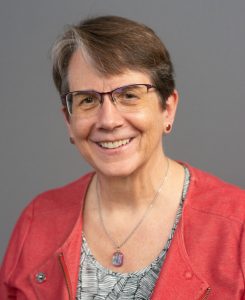 Judy Brewer: a woman with dark hair, glasses, wearing a necklace and coral colored top