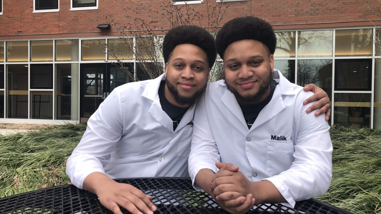 Twin brothers in lab coats sitting next to each other outside