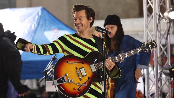 Harry Styles smiling while holding his guitar in a stripped jumpsuit on the Today Show. 