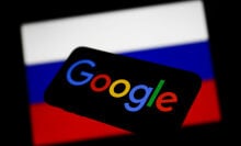 Google logo displayed on a phone screen and Russian flag displayed on a screen in the background.