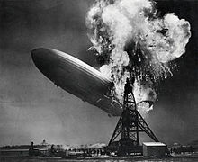 A black and white photograph of an airship near a mooring mast exploding at its stern.