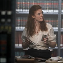 A woman stands in a library holding photos in one hand and a cassette tape in the other.