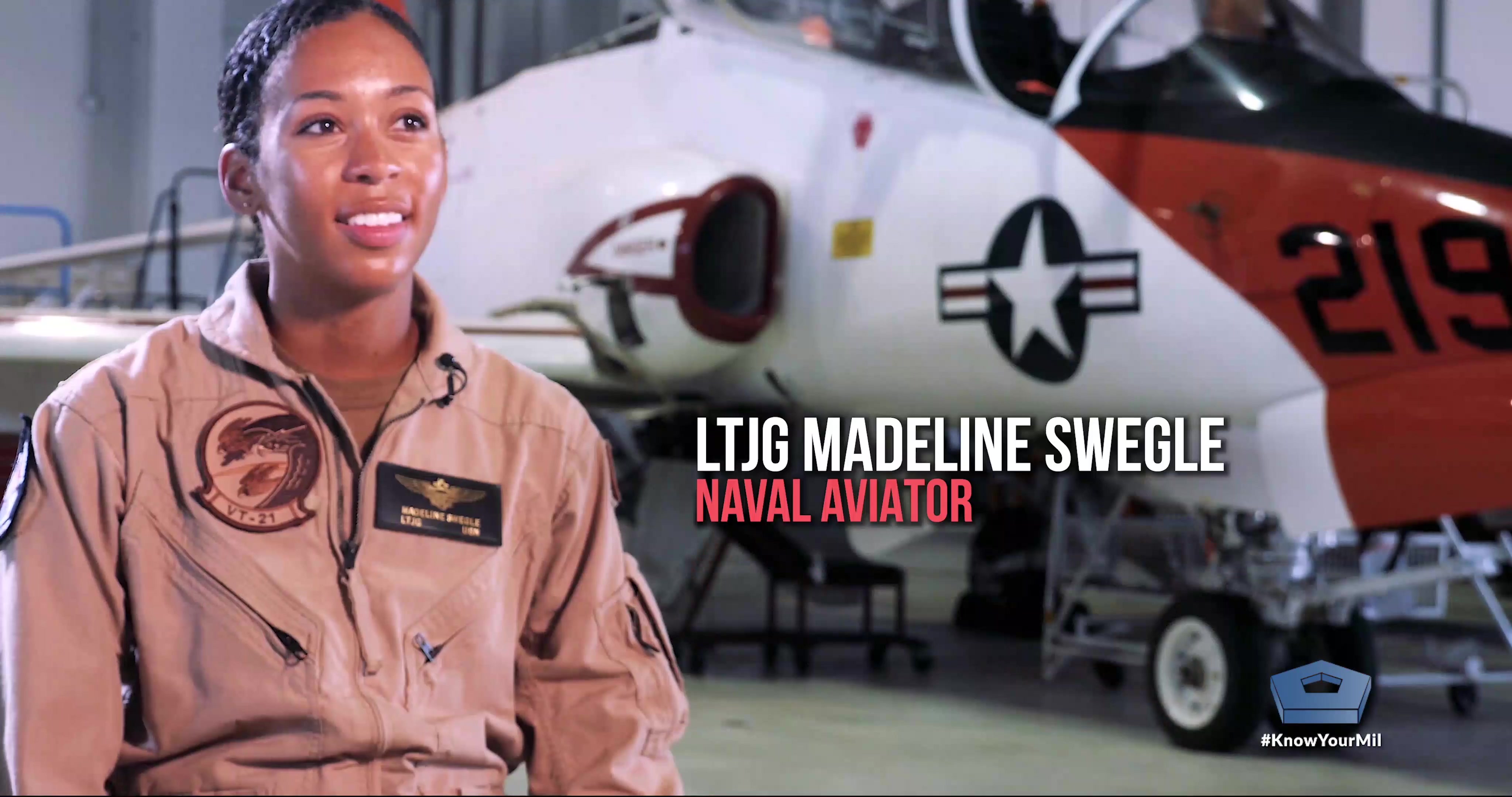 A sailor sitting in front of an aircraft in a hangar speaks. 