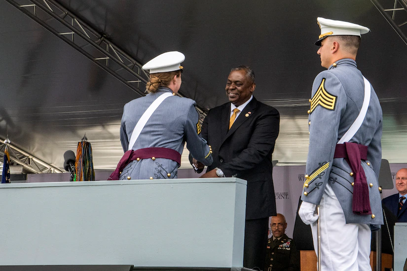 Defense Secretary Austin smiles and shakes hands with a U.S. Military Academy cadet on a podium.