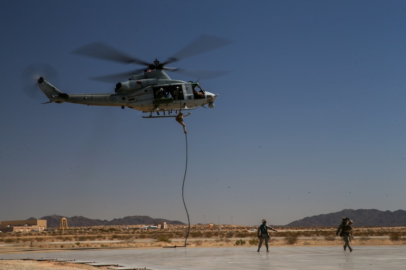 A Marine hands from a rope attached to a hovering helicopter as two others walk on a flightline below.