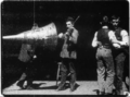 Black-and-white film screenshot of a man (at center) playing a violin while facing a large cone. An obscured man is seen behind the cone. Two men at the right are dancing with each other.