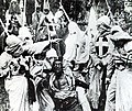 A black-and-white screenshot of several men in white robes and hoods surrounding an African-American man (a white actor using "blackface"). The men's robe have crosses on them and in the background other men are on top of horses.