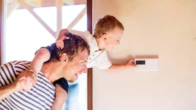 A father carrying his son on his shoulders while the child adjusts the temperature using a thermostat