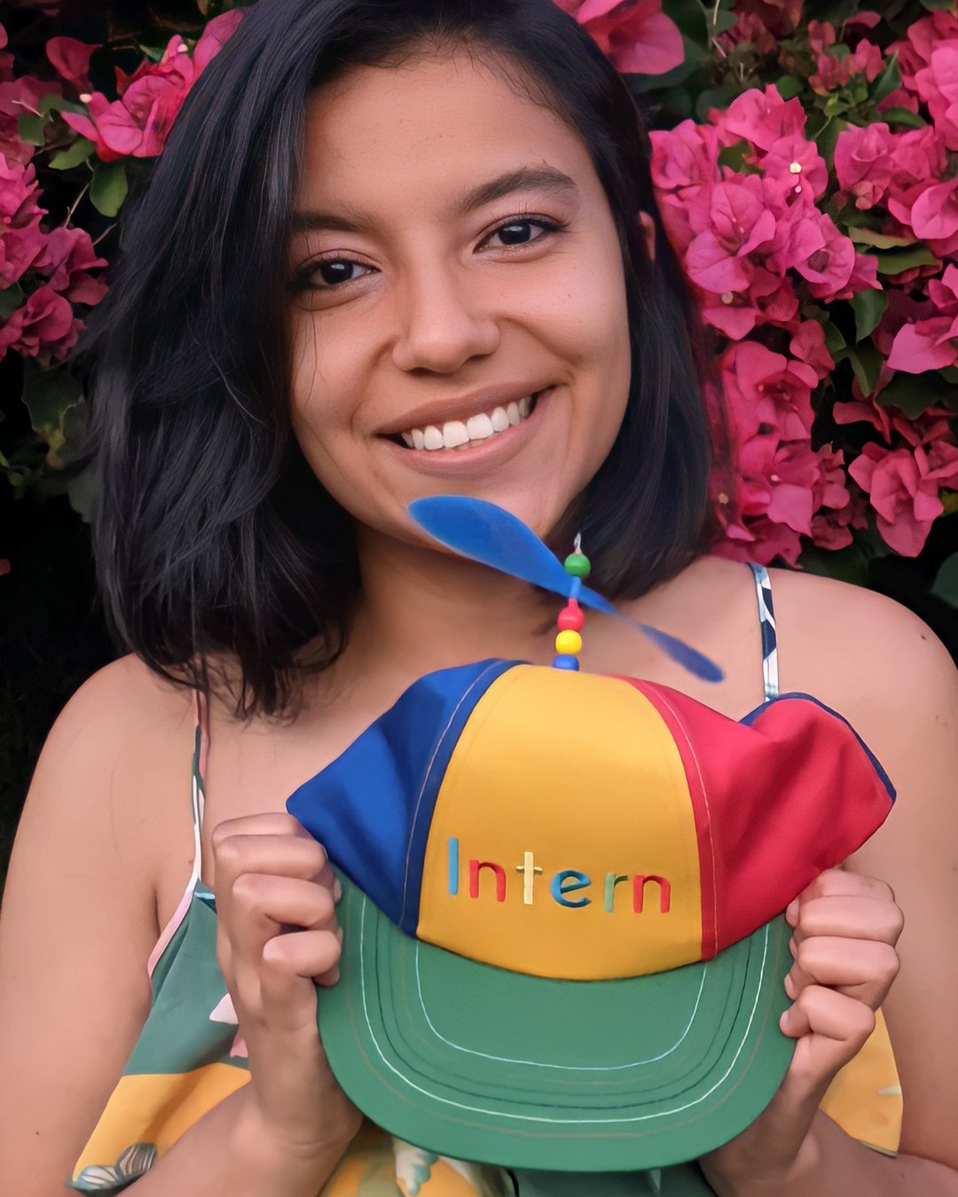 Micka holding a red, yellow, green, and blue Intern hat while smiling in front of pink flowers.
