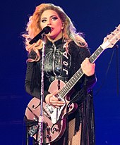 Lady Gaga standing behind a microphone stand with a pink guitar in her hands, wearing black leather fringe.