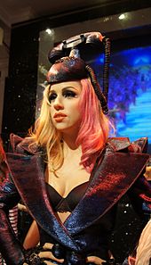 A realistic mannequin of a pale-skinned woman with blonde hair wearing a hat in the design of an old-fashioned telephone.