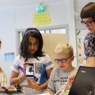 A teacher is conversing with four schoolchildren in an exercise where a book, a laptop and a mobile phone are all in use.