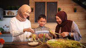 Two women in traditional Malaysian Muslim clothing and a boy stand at a kitchen counter weaving palm leave parcels.