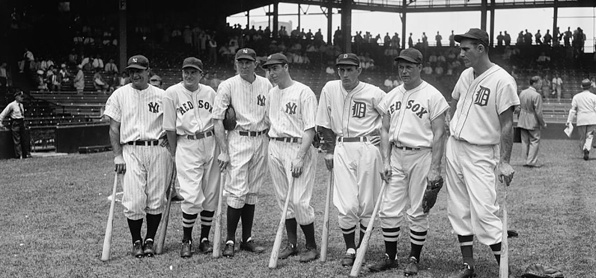 Image of men in baseball uniforms from a variety of teams posing in a iine on a ballfield holding baseball bats