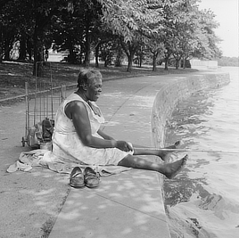 Smiling  woman sitting on the ground with a fishing pole and her bare feet dangling over the water.