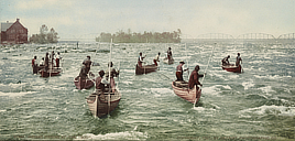 Pairs of men in several canoes that face into the water rapids.