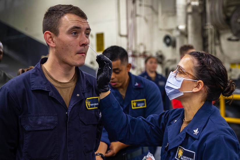 A service member wearing a face mask and gloves holds a nasal swab while administering a COVID-19 test to another service member.