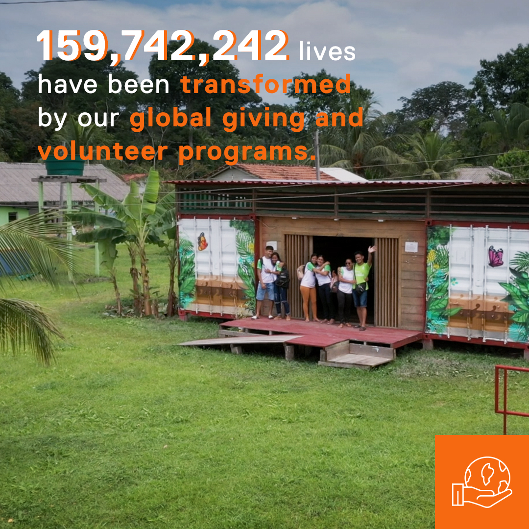 An image of children standing outside one of the Dell Technologies Solar-powered Learning Labs. The Learning Lab is surrounded by very green grass and palm trees. 

Text overlay on the image reads: 159,742,242 lives have been transformed by our global giving and volunteer programs.

An orange icon overlay of a hand holding the earth appears in the lower right corner. 
