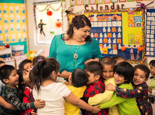 Smiling woman in a school classroom surrounded by a group of young children.