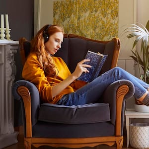 woman with headphones on reading on chair next to a fireplace