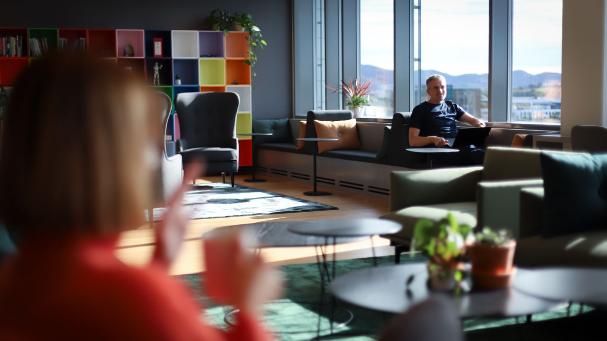 A man is sitting with his feet up in a colorful office communal area talking to a woman seen blurred in the foreground.