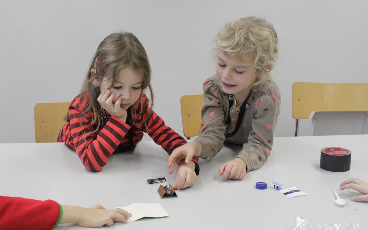 Zwei Mädchen sitzen am Tisch und basteln einen kleinen Roboter aus einem Motor, einem Zahnbürstenkopf und einigen anderen Utensilien.